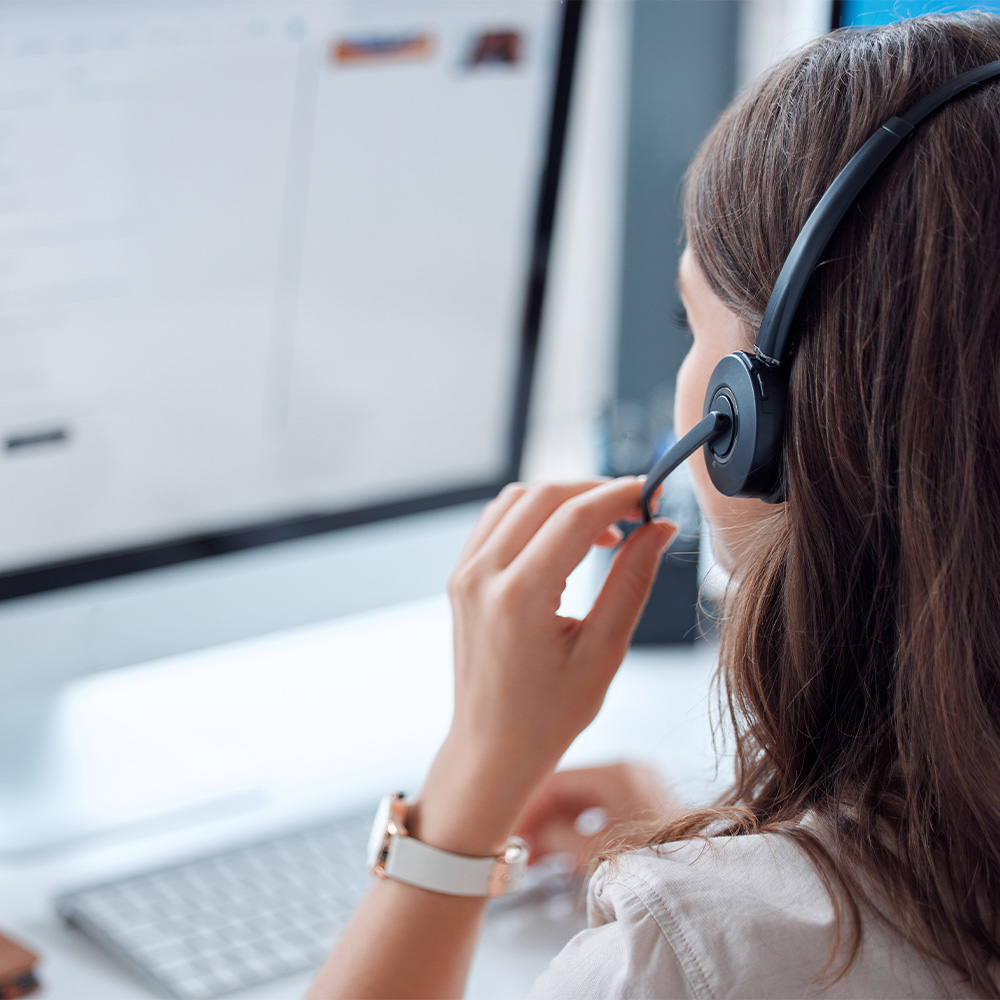 Frau mit Headset sitzt vor Computerbildschirm und hält das Mikrofon nahe am Mund