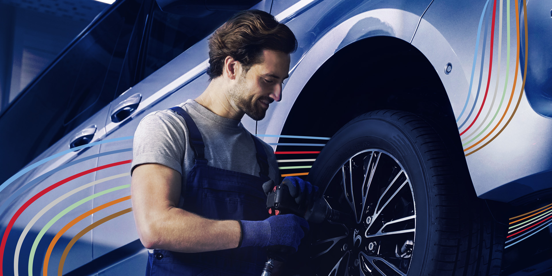 Ein Mechaniker in blauer Arbeitskleidung und Handschuhen bearbeitet mit einem Elektrowerkzeug einen glänzenden Autoreifen. Das Auto hat eine Metallic-Lackierung mit bunten Streifen an den Seiten.